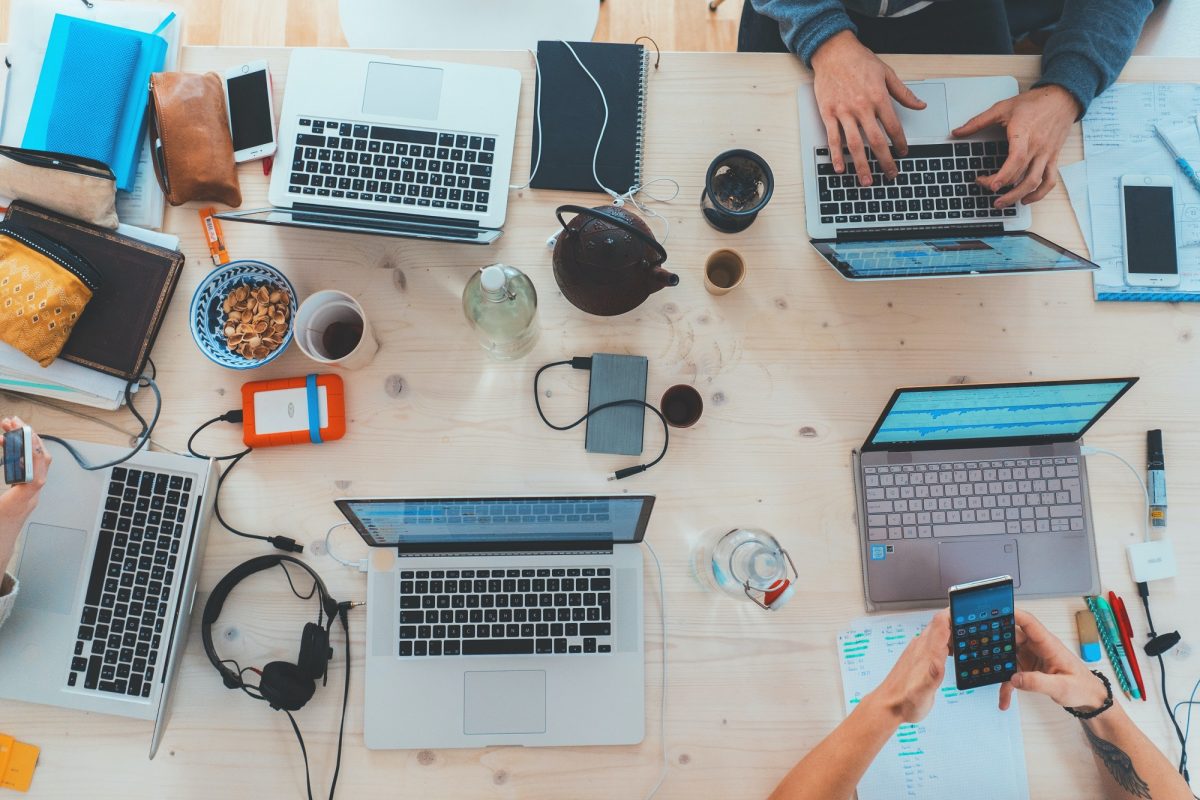 A table is covered with open laptops, water bottles, pencil cases, and other various items as people sit in front of the laptops