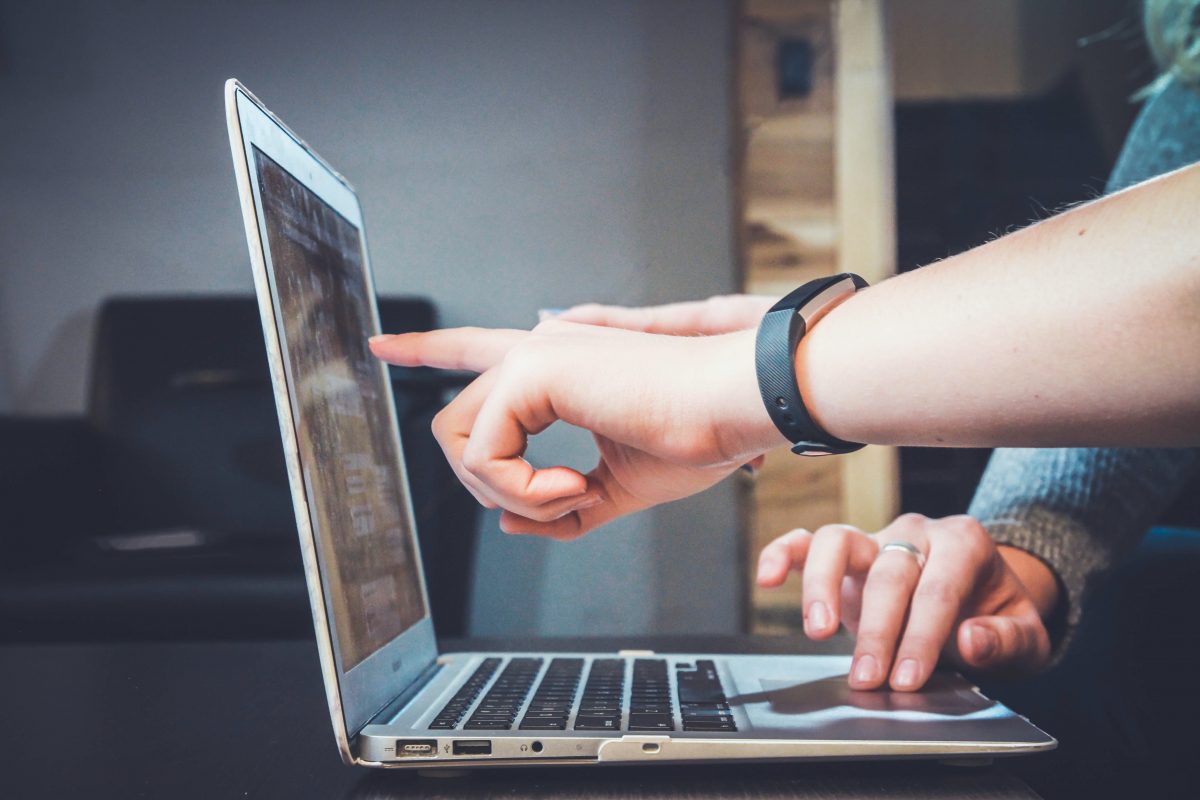 A hand is pointing at a laptop screen while another set of hands are using the laptop's trackpad